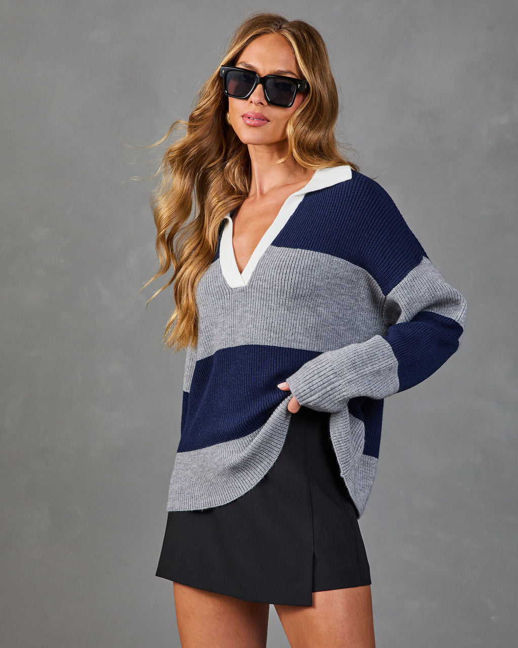 Woman wearing oversized navy and gray striped knit polo sweater with white collar, black mini skirt, and square sunglasses
