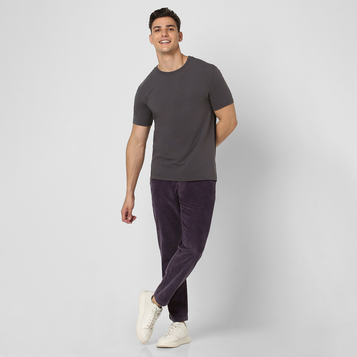 A man stands against a light gray background, smiling and posing with one foot raised and hands behind his back. He wears a dark gray short-sleeve tee, Corduroy Easy Pant in dark purple cotton, and white sneakers.